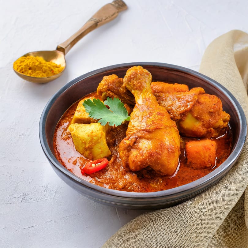 Chicken curry with potatoes and red chili in a dark bowl, with a spoon of turmeric powder.