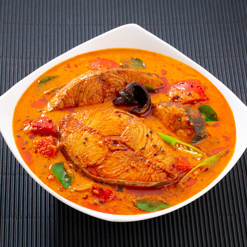 Indian fish curry with red chili, green peppers, and curry leaves in a white bowl.